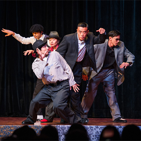Five performers grouped close and holding poses on stage in front of an audience.